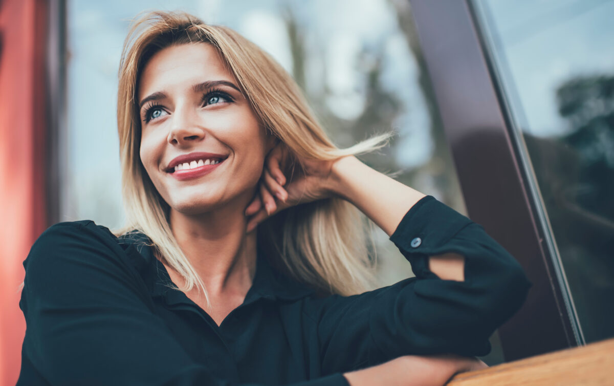 Happy hipster girl with perfect white teeth sitting on bench and laughing during free time, positive cheerful female teenager feeling good and gladness on urban setting, concept of candid smile